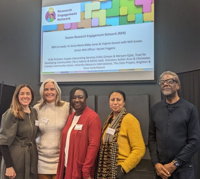 A group of five people smiling, underneath a projected slide showing information about the Sussex Research Engagement Network (REN)