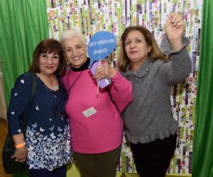 Three women standing in front of the SIS green backdrop, one is holding a speech bubble with the words "Let's celebrate diversity"